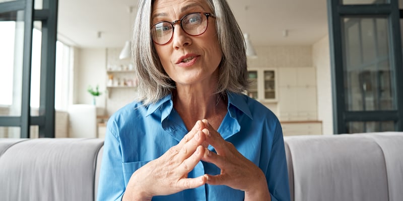 A senior woman wearing glasses is seated on a couch, talking about the menopause weight gain.