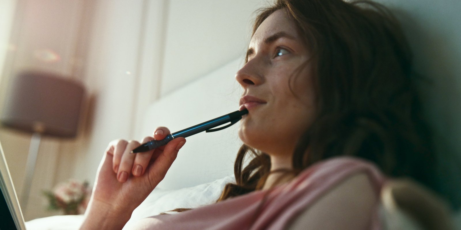Woman lying on the bed with a pen on her chin looking straight